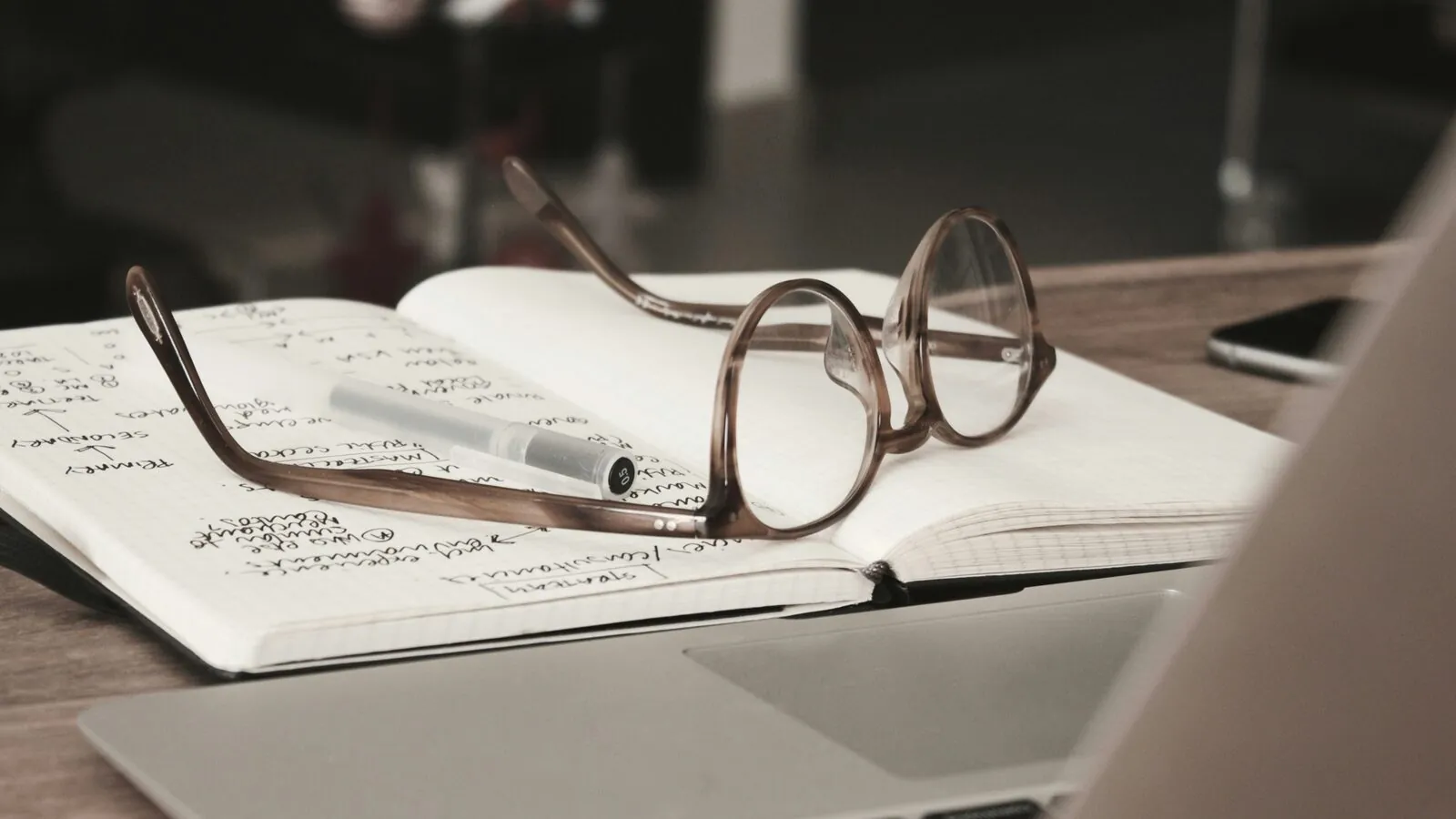 A pair of glasses and a pen resting on an open notebook of handwritten research notes beside a laptop