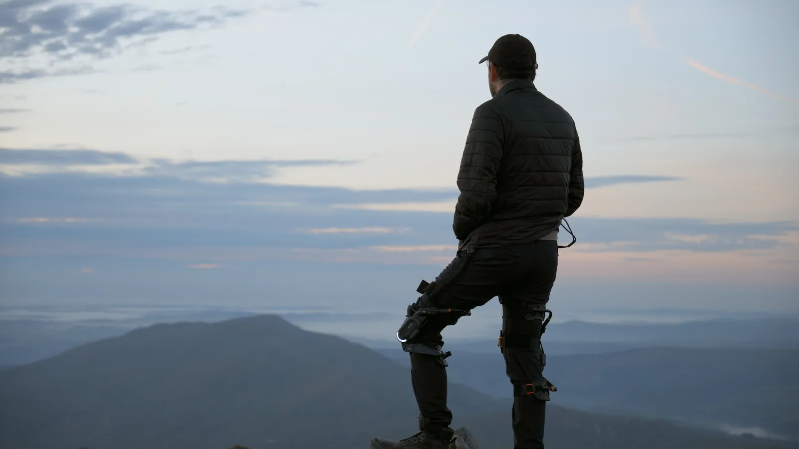 A man wearing a DNSYS Z1 exoskeleton on his legs stands on a rock looking out over misty mountains at dawn